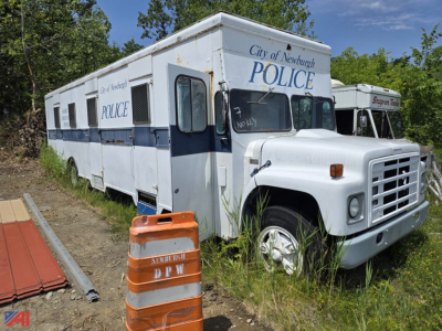 Mobile police station truck with City of Newburgh POLICE markings on white and blue exterior, side windows, and orange roadside barrier in foreground.