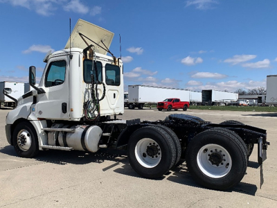 Freightliner Cascadia 126 tractor side view showing 3 axles with twin rear wheels and exposed engine bay area; white cab, no trailer, blue sky.