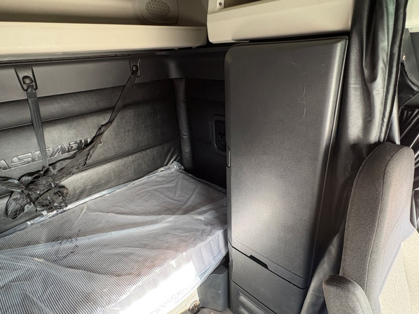 Freightliner Cascadia 126 sleeper cab interior showing storage cabinet and tarp-covered bed area, seat in foreground, gray upholstery.