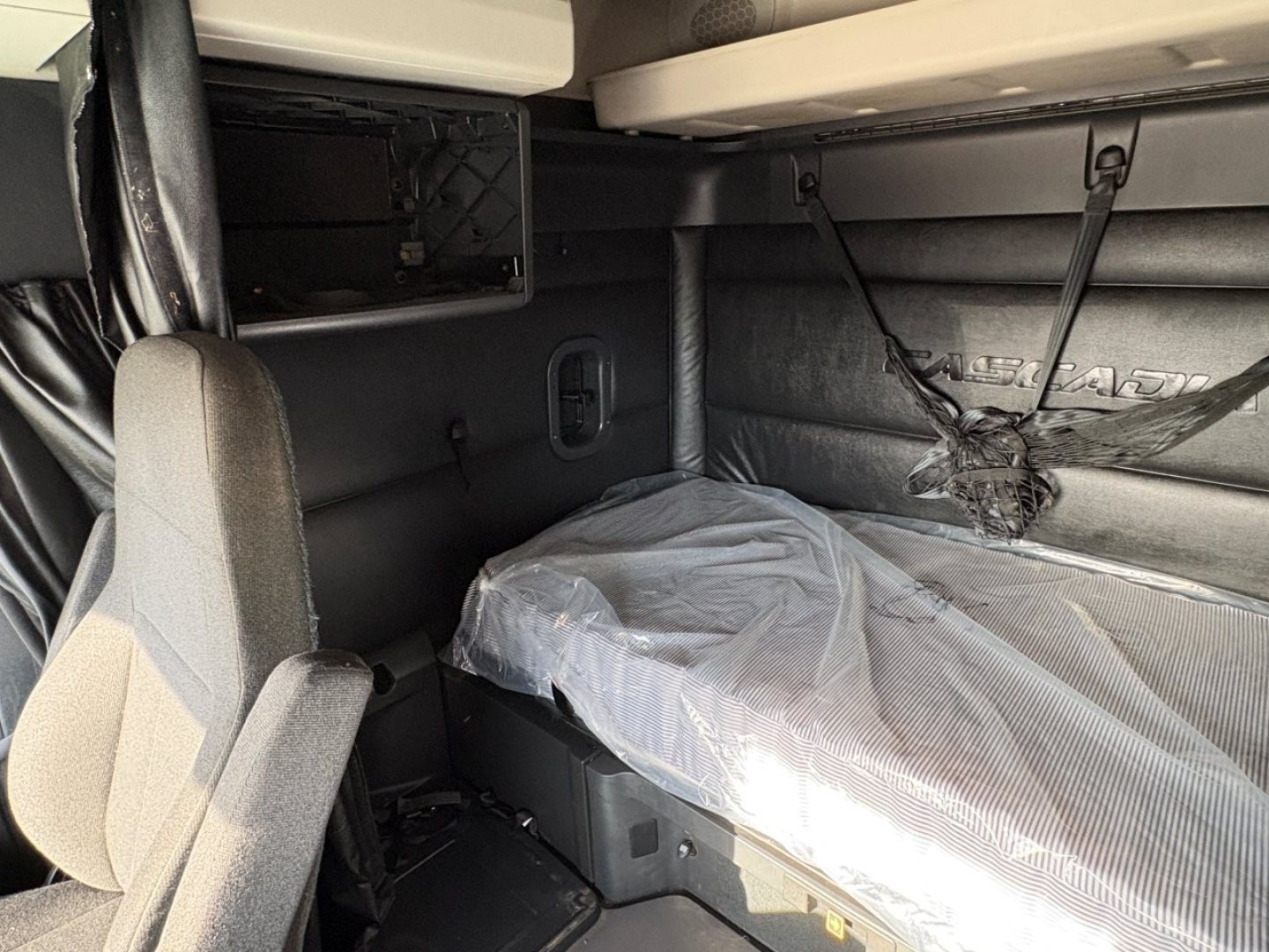 Interior of a Freightliner Cascadia sleeper cab showing bunk area with plastic-covered mattress, sidewall panels, and overhead cabinetry.