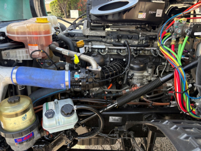 Truck engine bay of a 2023 Freightliner Cascadia 126 showing coolant reservoir, hoses, filter canister, battery wiring harness, and various fluid reservoirs.