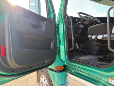 Cabin entry of a green Freightliner Cascadia tractor showing open driver door and dashboard area with steering wheel and pedals visible.