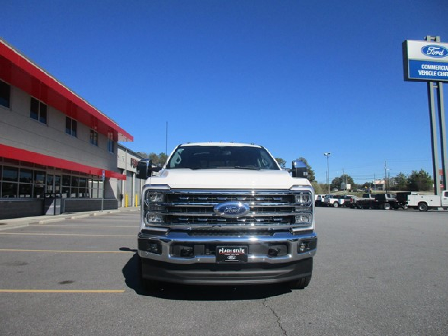 Front view of a white 2026 Ford F350 pickup truck in a dealership lot, showing grille, headlights, Ford emblem, chrome bumper, and dealership signage in background.