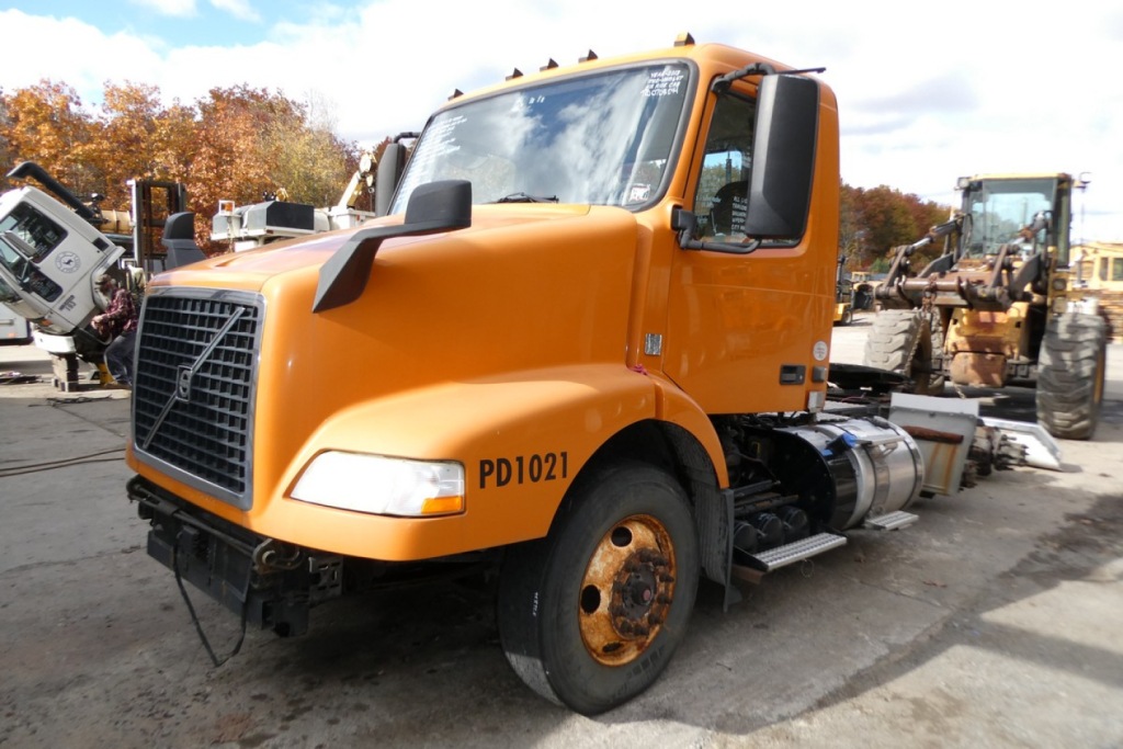 Orange Volvo VNM64T conventional day cab truck front-left view with PD1021 on fender and other construction equipment in background.