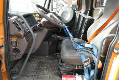 Interior of a Volvo VNM64T truck cab showing steering wheel, driver seat with blue seatbelt, dashboard controls, and protective floor mat.