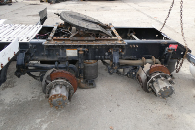 Truck chassis with rear axle components and brake drums exposed; hydraulic reservoir and mounting frame visible; rusty wheel hubs and fifth-wheel mounting plate.
