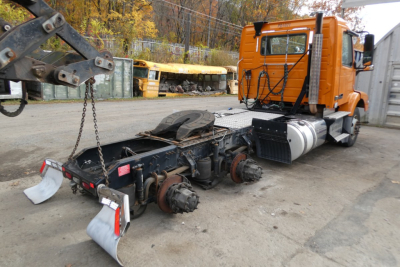 Volvo VNM64T truck chassis and cab in a yard showing orange cab and exposed rear drivetrain with axles and mud flaps.
