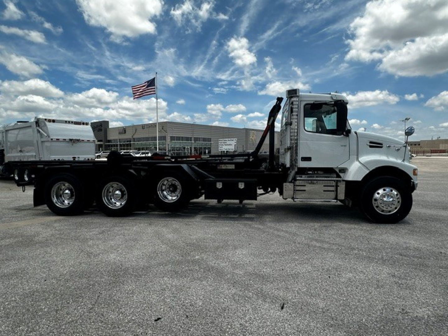 A white Volvo VHD roll-off truck in a parking lot with a flag in the background, long chassis with multiple axles and a roll-off bed.