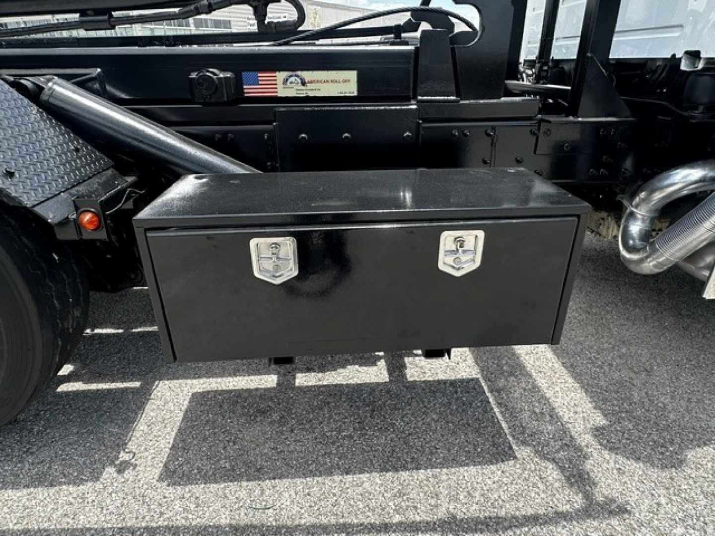 Truck chassis with a black rectangular toolbox mounted under the cab; two chrome latches on the toolbox; Volvo VHD chassis visible with metal steps and exhaust pipe; sticker with US flag on frame.