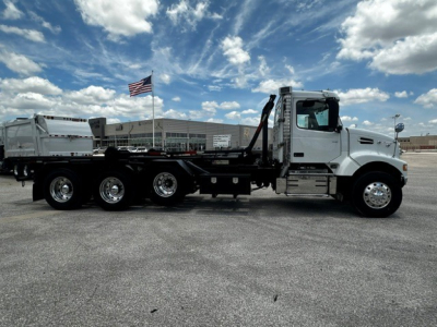 A white Volvo VHD roll-off truck in a parking lot with a flag in the background, long chassis with multiple axles and a roll-off bed.