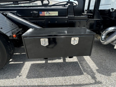 Truck chassis with a black rectangular toolbox mounted under the cab; two chrome latches on the toolbox; Volvo VHD chassis visible with metal steps and exhaust pipe; sticker with US flag on frame.