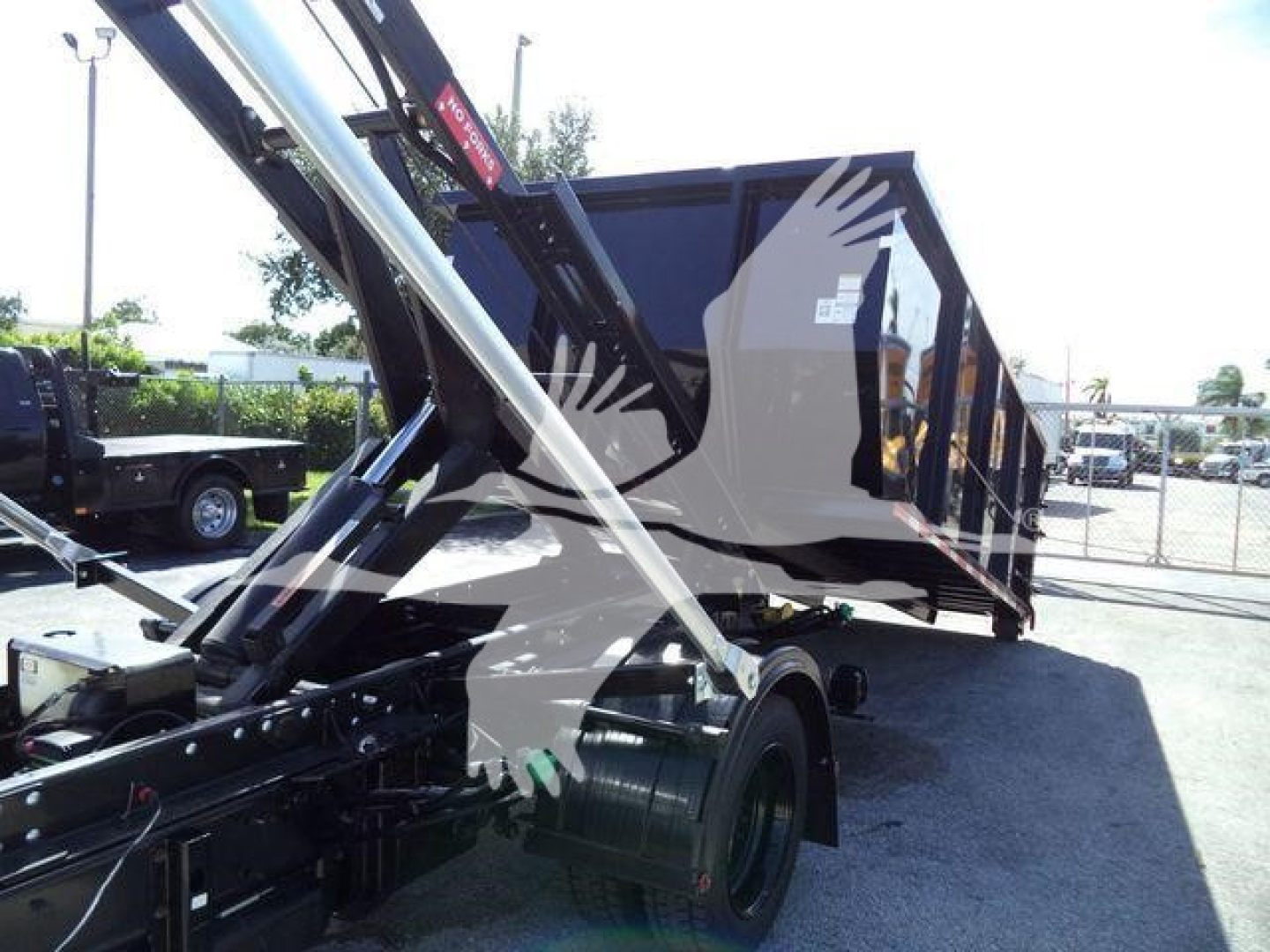 Truck bed and lift arm of a 2023 Chevrolet Silverado 6500HD roll-off truck with hydraulic arms and a dump bed”, showing partial side view of a black/dark gray vehicle chassis and container.