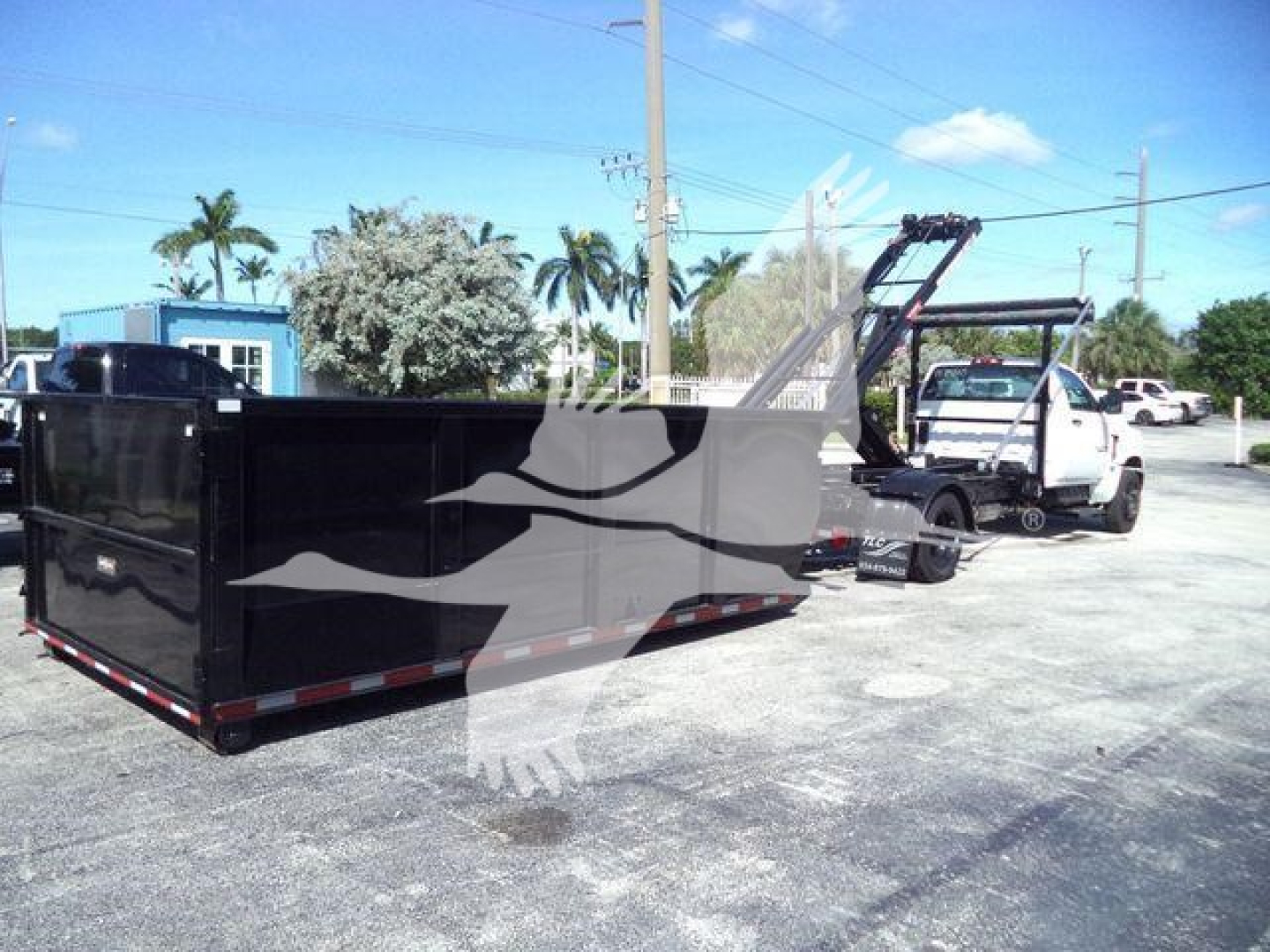 Truck in a lot with a black open-top dumpster trailer and a white tow truck in background. Clear sky with palm trees.