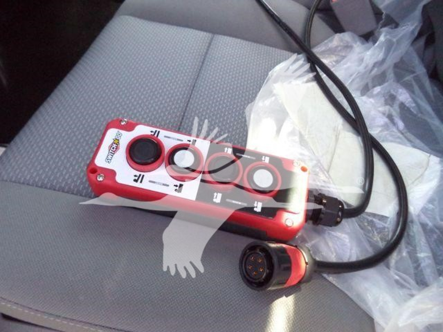 Control pendant with red housing and multiple buttons on vehicle seat; circular connector at end of cable.