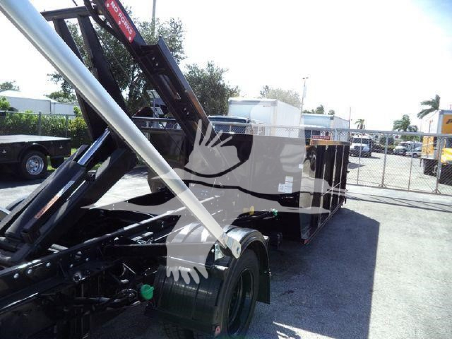 Truck chassis and roll-off truck bed with hydraulic hoist in a dealership lot, side view of rear wheels and lifting mechanism.