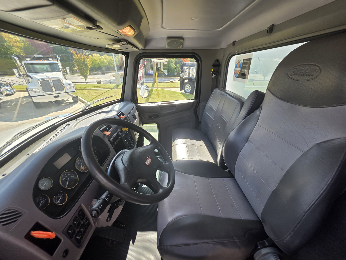 Peterbilt 337 interior cab view showing steering wheel, dashboard gauges, driver seat and passenger seat with grey vinyl. side window and view of other trucks outside.