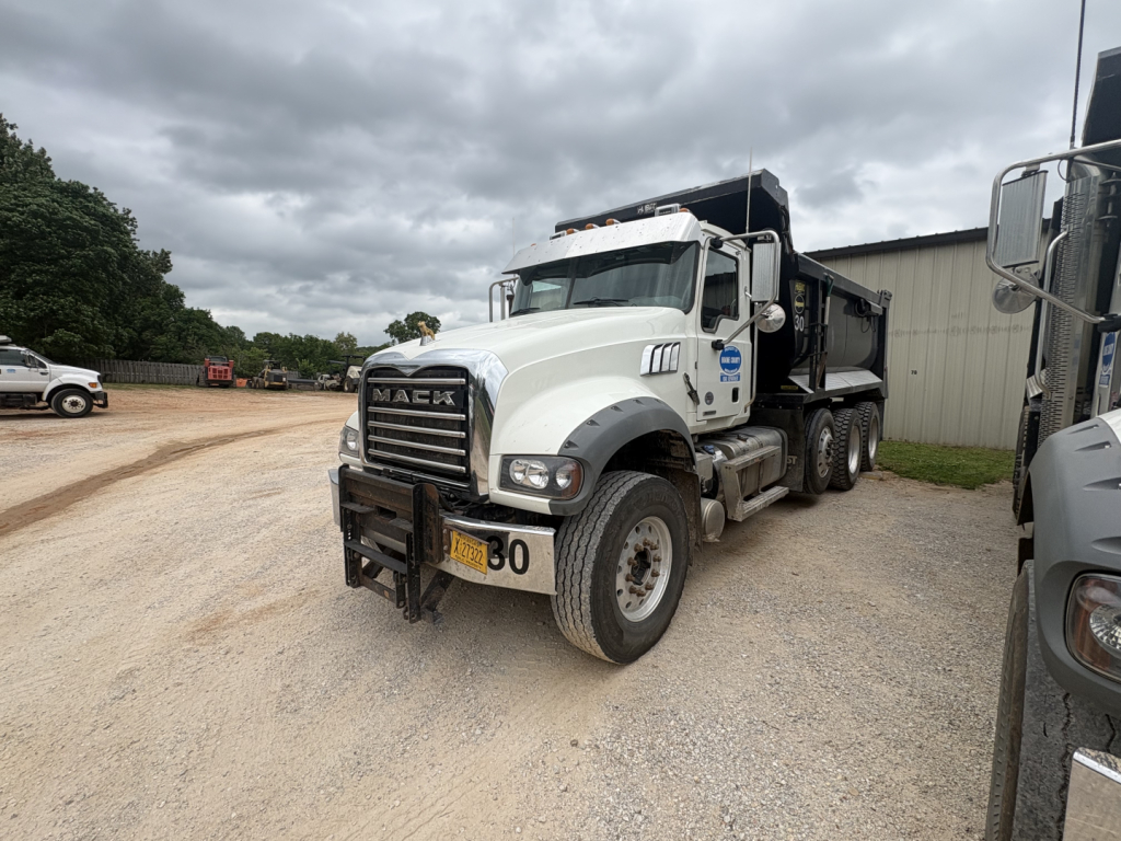Mack GR84F dump truck in a gravel lot, front three-quarter view showing chrome grille, bumper with numbered sign, and dump bed.