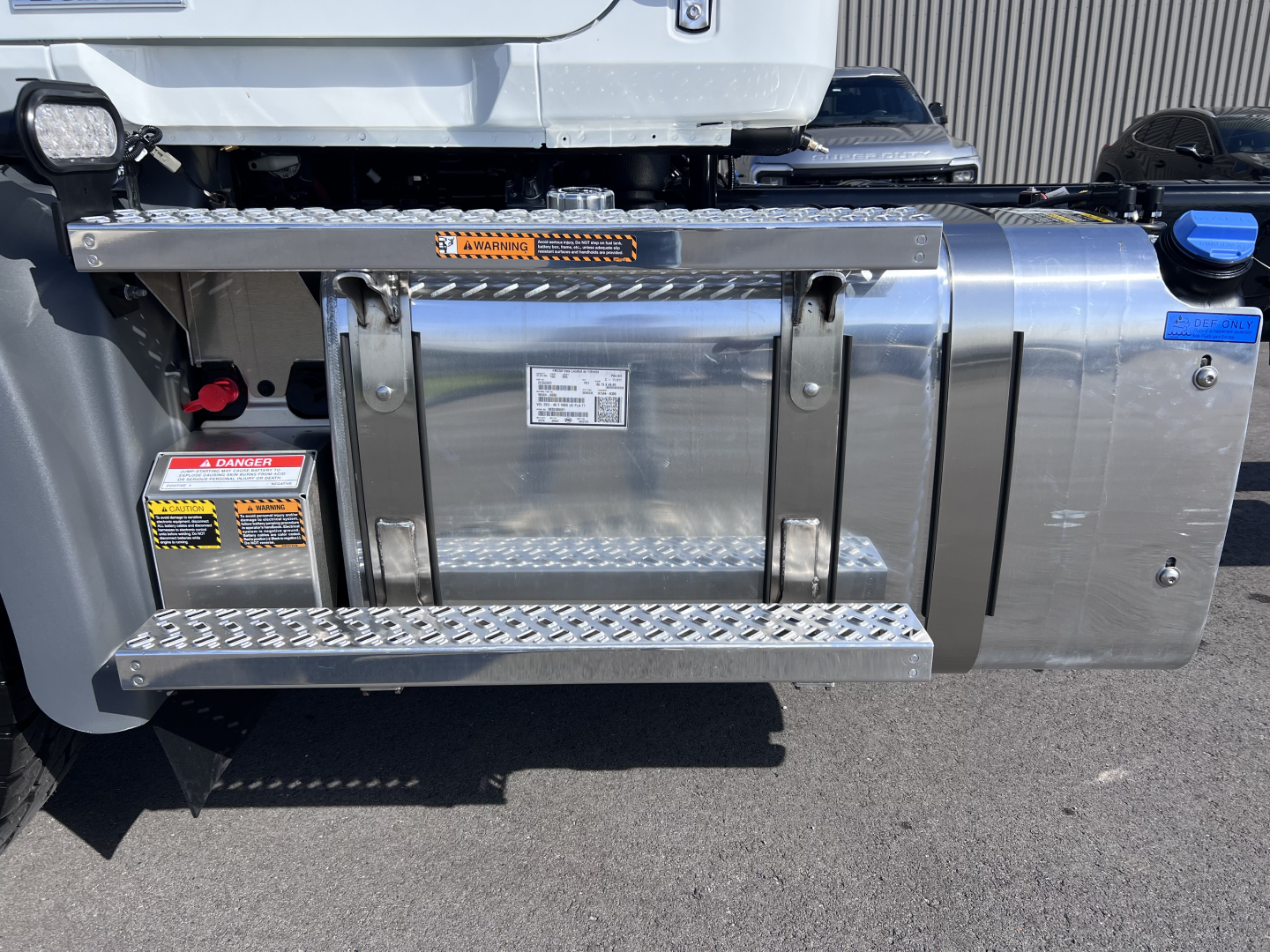 Truck fuel tank area on Mack GR64B cab and chassis showing polished metal fuel tank, step plates, safety labels, and hoses.
