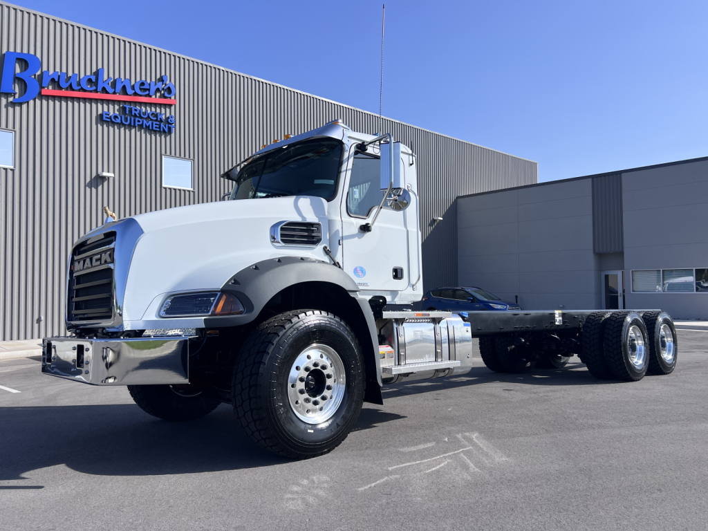 Mack GR64B cab chassis truck in a dealership lot with white cab and dual rear axles, chrome front bumper, and logo on building in background.