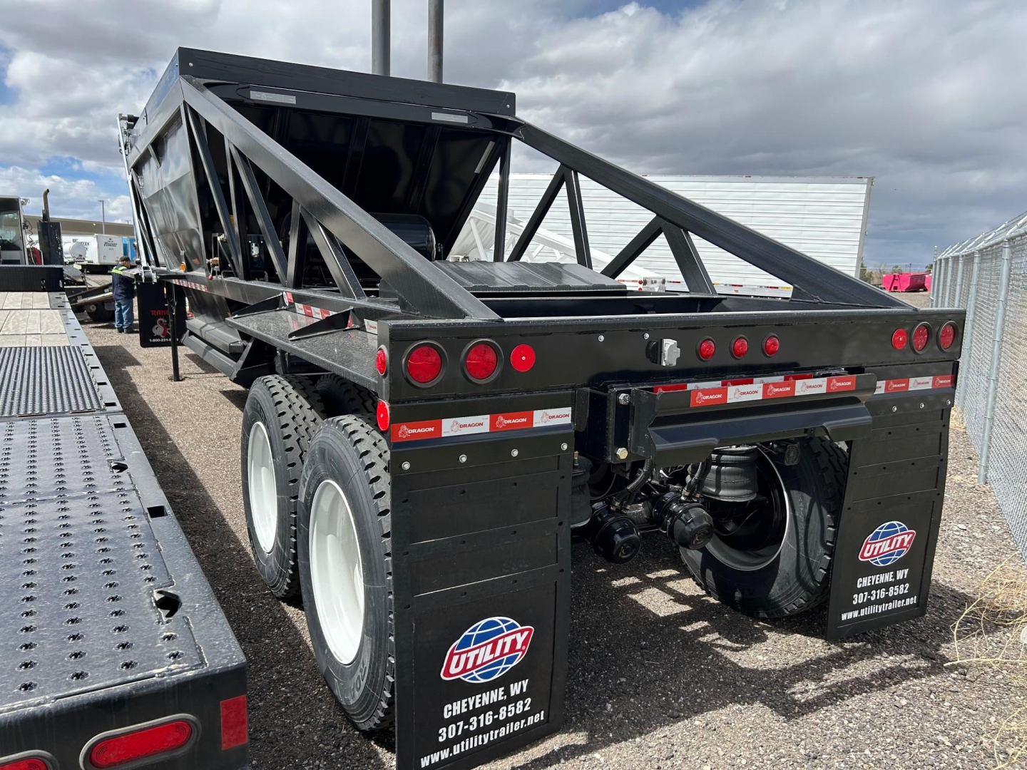 Dump trailer showing rear view with dual rear axles and black steel frame. Visible Utility logos, reflective tape, lights, and rear hitch area.