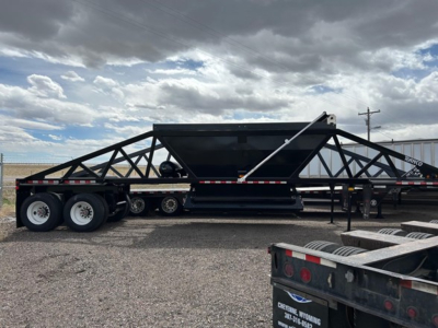 Large black dump trailer frame with truss-like steel supports and a central hopper, three-axle tandem rows on a gravel lot, blue sky with clouds.