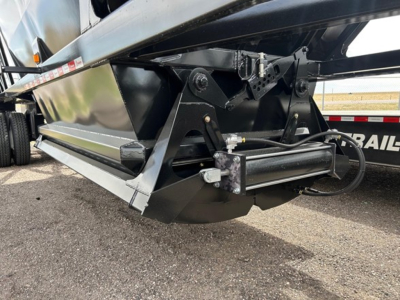 Dump trailer undercarriage showing hitch mechanism and hydraulic components with black metal frame and angled skid plate.