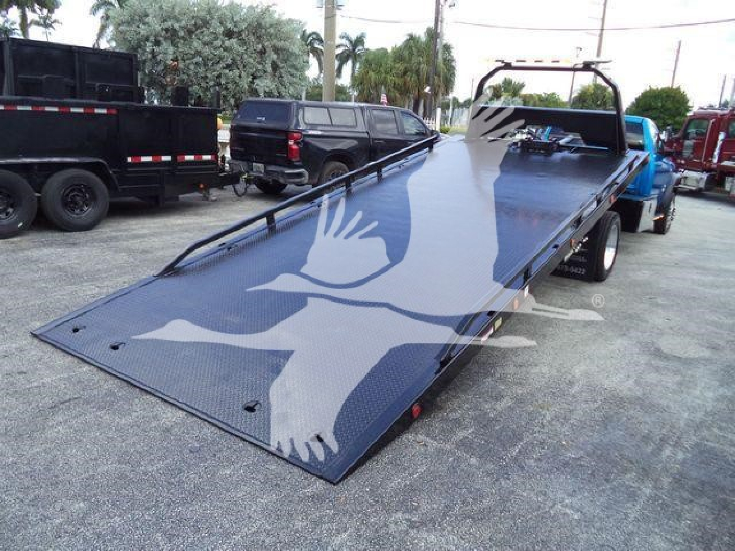 Ram 5500 Rollback truck showing a long metal tilt bed with black textured surface and wheel wells; several trucks parked nearby.