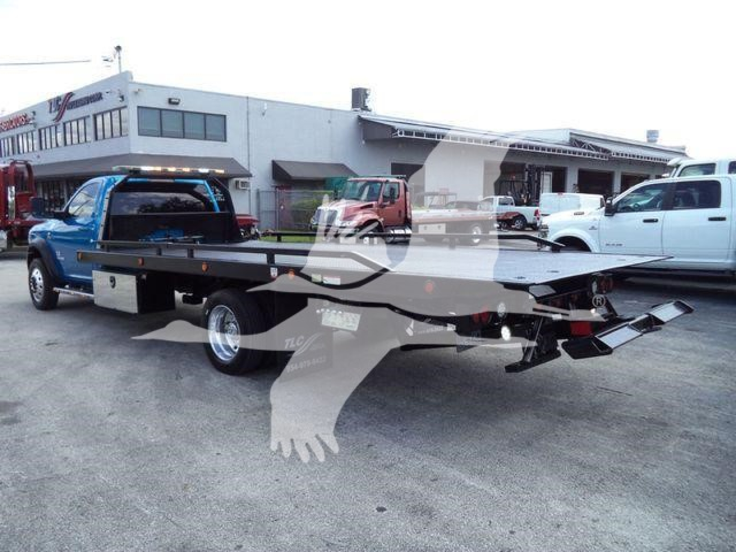 Ram 5500 rollback tow truck with flatbed showing rear bed and hoist, blue cab, dealership lot in background.