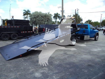 Ram 5500 rollback tow truck with a tilted flatbed, blue pickup in background, dealership lot scene.