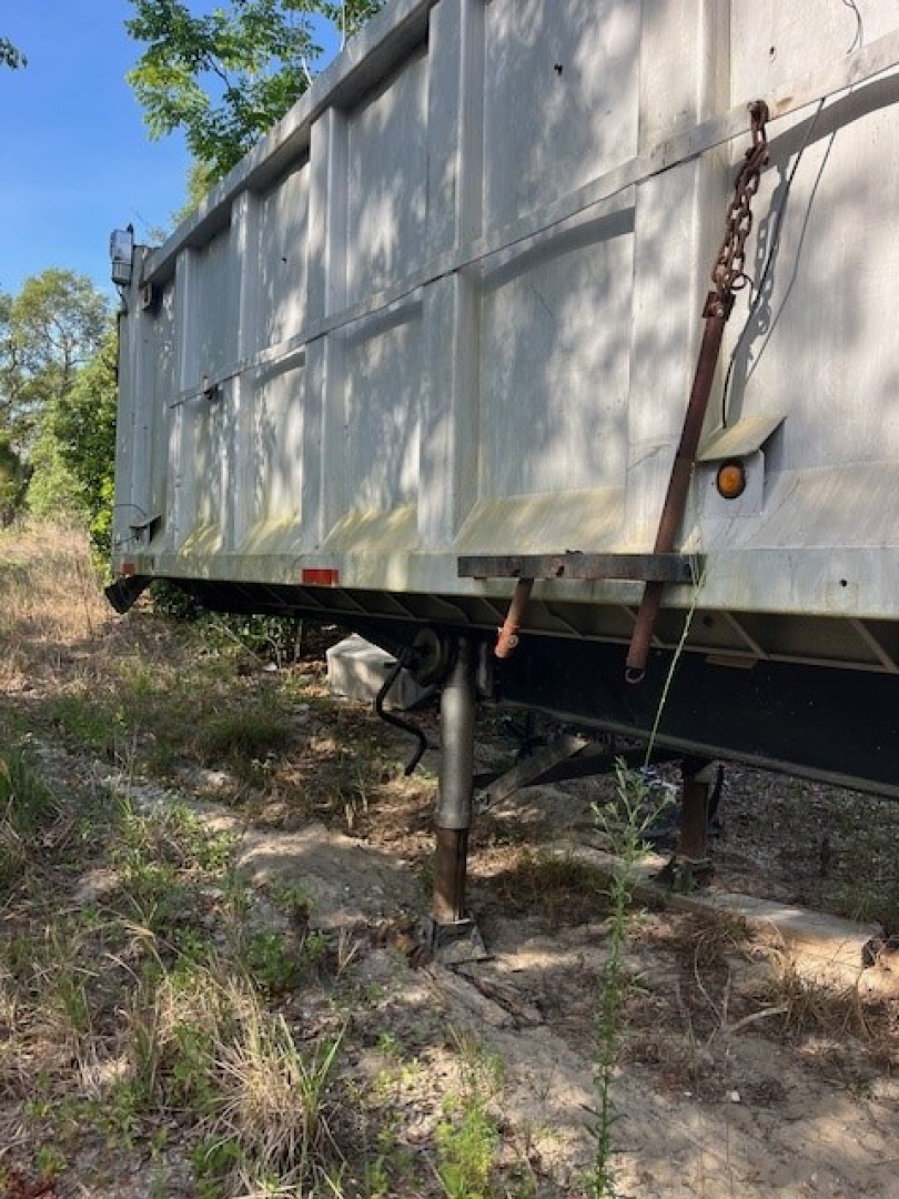 Dump trailer undercarriage showing support jack and hitch assembly with chain and rustic terrain.