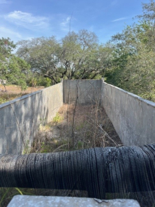 Dump trailer interior showing metal walls of a long rectangular bed with overgrown vegetation; view from front of trailer with black flexible pipe in foreground.