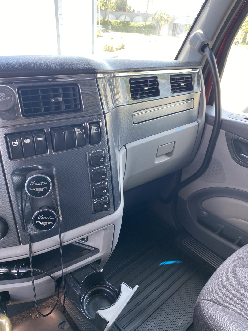 Truck dashboard and front passenger area of a 2015 Peterbilt 567 conventional sleeper showing dashboard controls, vents, cup holder, and beige interior.