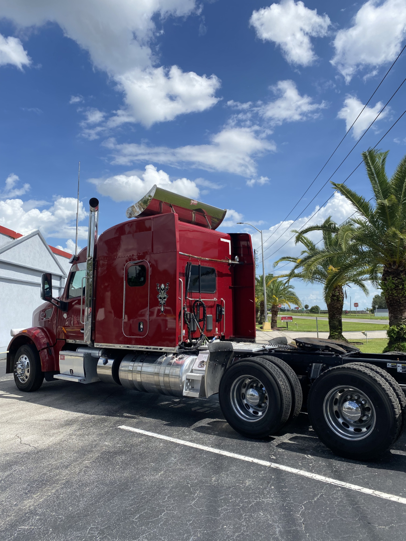 Peterbilt 567 conventional sleeper truck in a parking lot showing red cab and rear dual wheels with chrome fuel tanks.