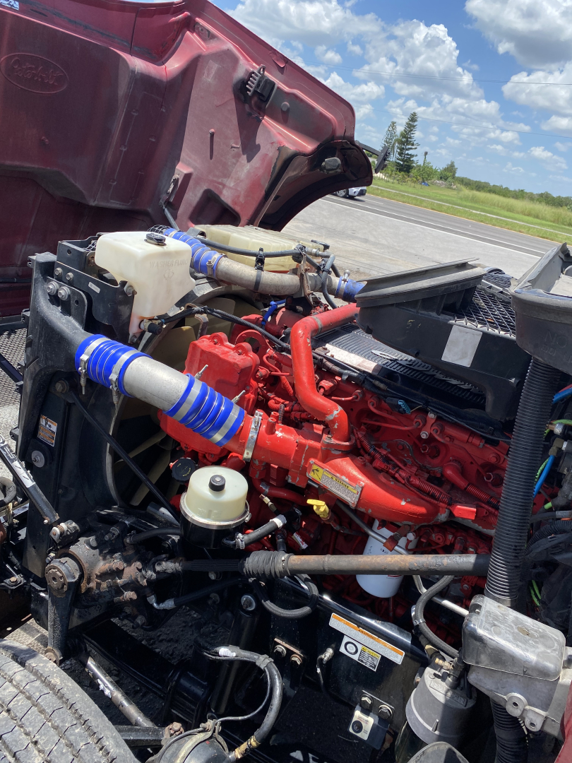 Peterbilt 567 engine bay showing red engine with blue silicone hoses, white coolant reservoir, black radiator, and various components.