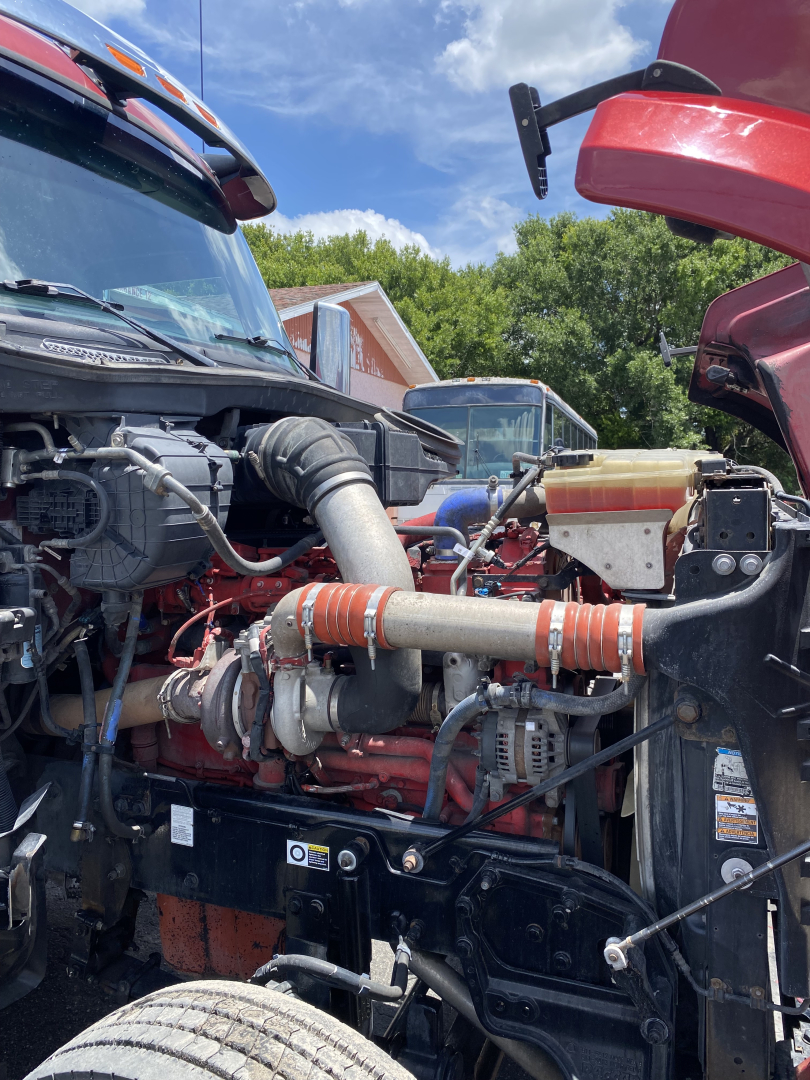 Peterbilt 567 engine bay view showing large intake tube, red engine block, hoses, and coolant reservoir”,