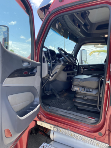 Truck cab interior of a 2015 Peterbilt 567 conventional sleeper, driver seat, steering wheel, dashboard, and entry door visible from open cab doorway.