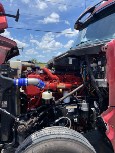Truck engine bay showing red diesel engine with blue booted hose, various cables, and in-service components under the hood of a 2015 Peterbilt 567 conventional sleeper truck.