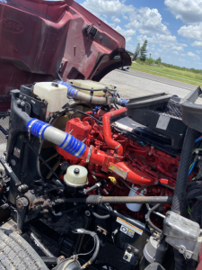 Peterbilt 567 engine bay showing red engine with blue silicone hoses, white coolant reservoir, black radiator, and various components.