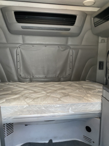 Interior sleeper cab of a 2015 Peterbilt 567 showing bunk bed with mattress, side storage pocket, and overhead vent; light gray fabric and panel surrounds.