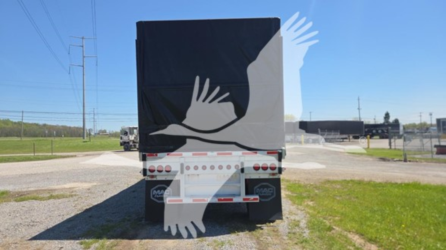 Rear view of a curtain-side trailer with black and white graphic of hands. Tail lights visible, bumper with truck license plate area, and a rural roadway in background.
