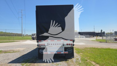 Rear view of a curtain-side trailer with black and white graphic of hands. Tail lights visible, bumper with truck license plate area, and a rural roadway in background.
