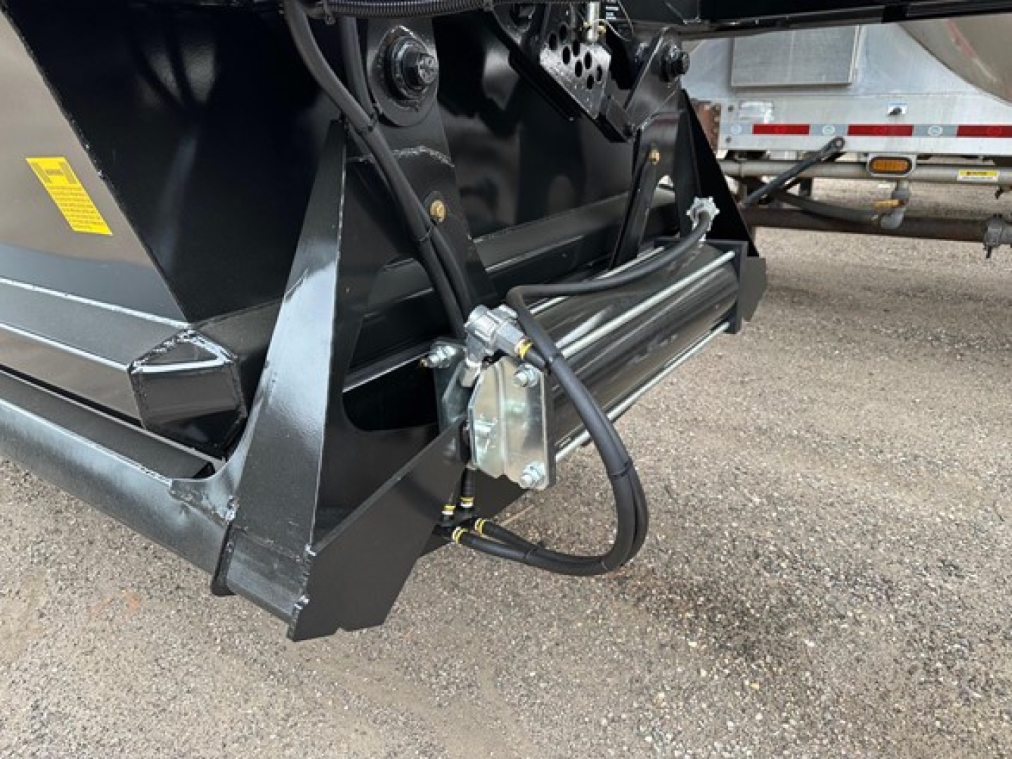 Dump trailer hitch area showing hydraulic hose and actuator assembly on a black metal frame, with a silver mounting bracket and hydraulic line connected to the trailer.