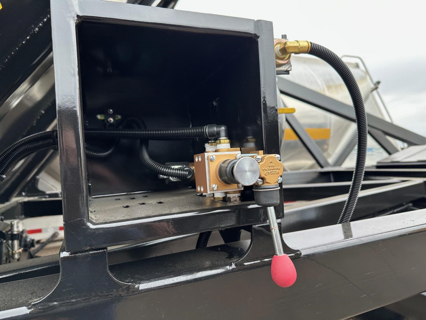 Close-up of a hydraulic control valve assembly mounted in a metal housing on a dump trailer, with a red-tipped lever and label on the valve.