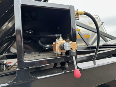 Dump trailer component showing hydraulic control valve assembly inside a square metal housing with hoses and a red-topped lever handle.