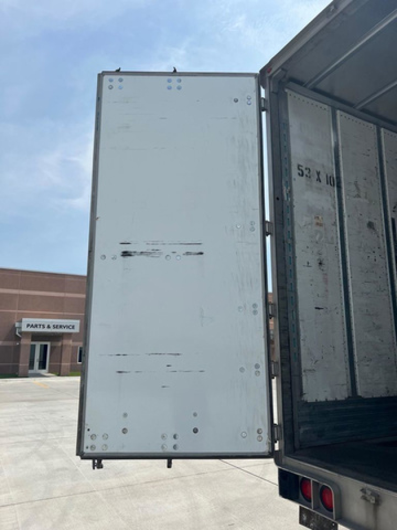 Rear of a utility van trailer with open doors showing interior metal panels and riveted edges; some wheel of a building in background.