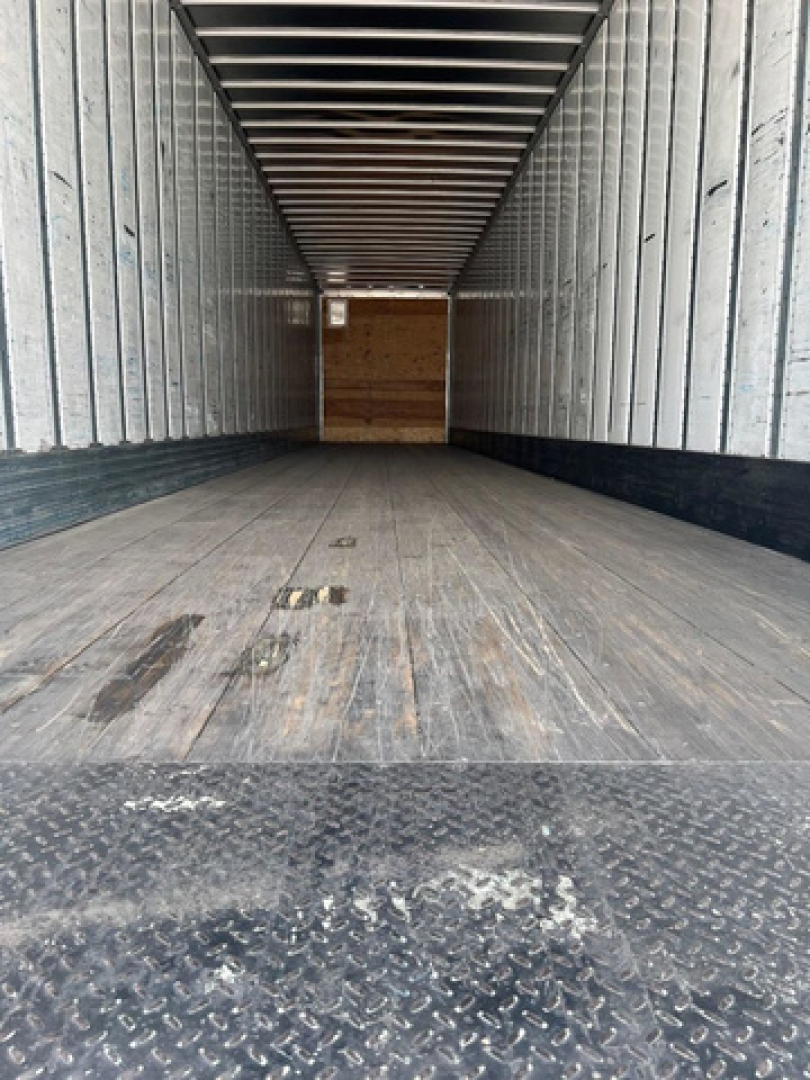 Trailer interior showing empty flatbed with wooden floor and metal walls, viewed from rear entry toward cab at far end.