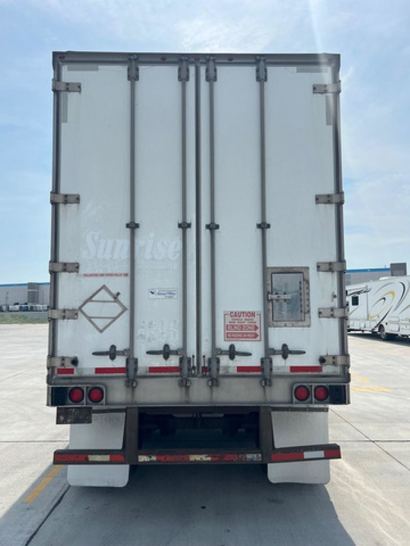 Rear view of a white utility van trailer with double rear doors, taillights, and warning labels, parked on a lot.