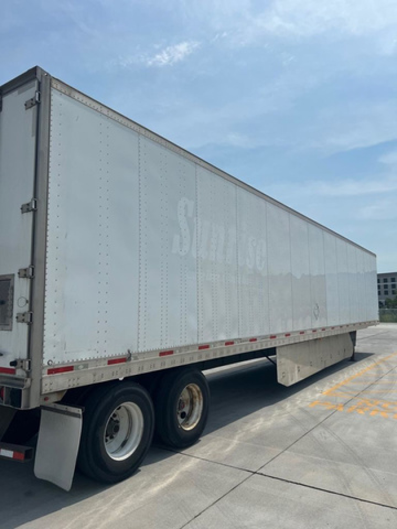 Trailer side view showing a long white utility van trailer with dual rear axles and reflective tape along the bottom edge.