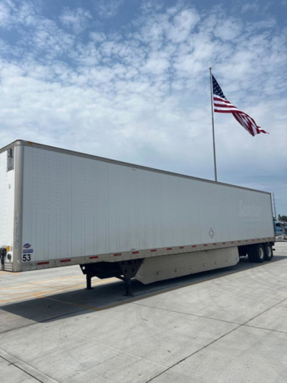 Utility van trailer with a white box, landing gear visible, and an American flag in the background. Side of trailer shows no readable branding.