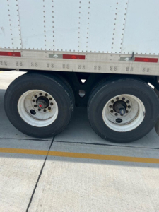 Trailer side showing dual rear wheels of a utility trailer with white rims and black tires; visible riveted side panel with small red/white reflectors; yellow painted line on pavement